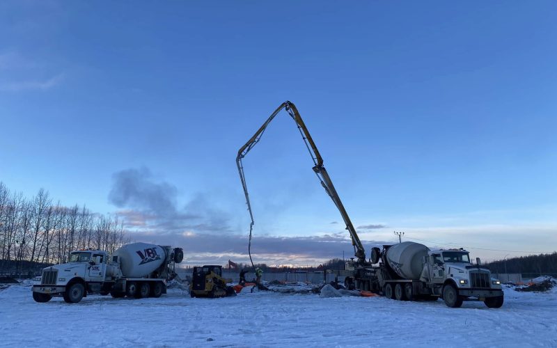 Concrete pumping trucks operating on a snowy construction site in Alaska at sunset