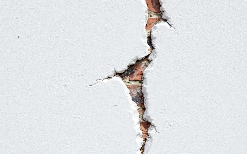 Close-up of a large crack in a white concrete wall exposing red bricks underneath